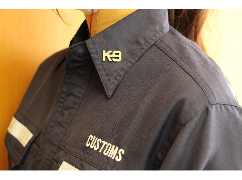 麻薬探知犬ハンドラーの制服襟元には、K-9のバッジが。K-9は英語で「犬」という意味。現在では、人々のために働く犬の代名詞として使われている